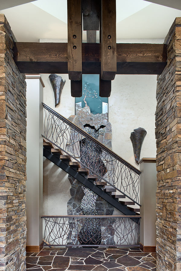 Walnut and steel floating staircase with metal stair rail of interwoven branches in front of double-height water feature with tile mosaic, infused metal and art glass